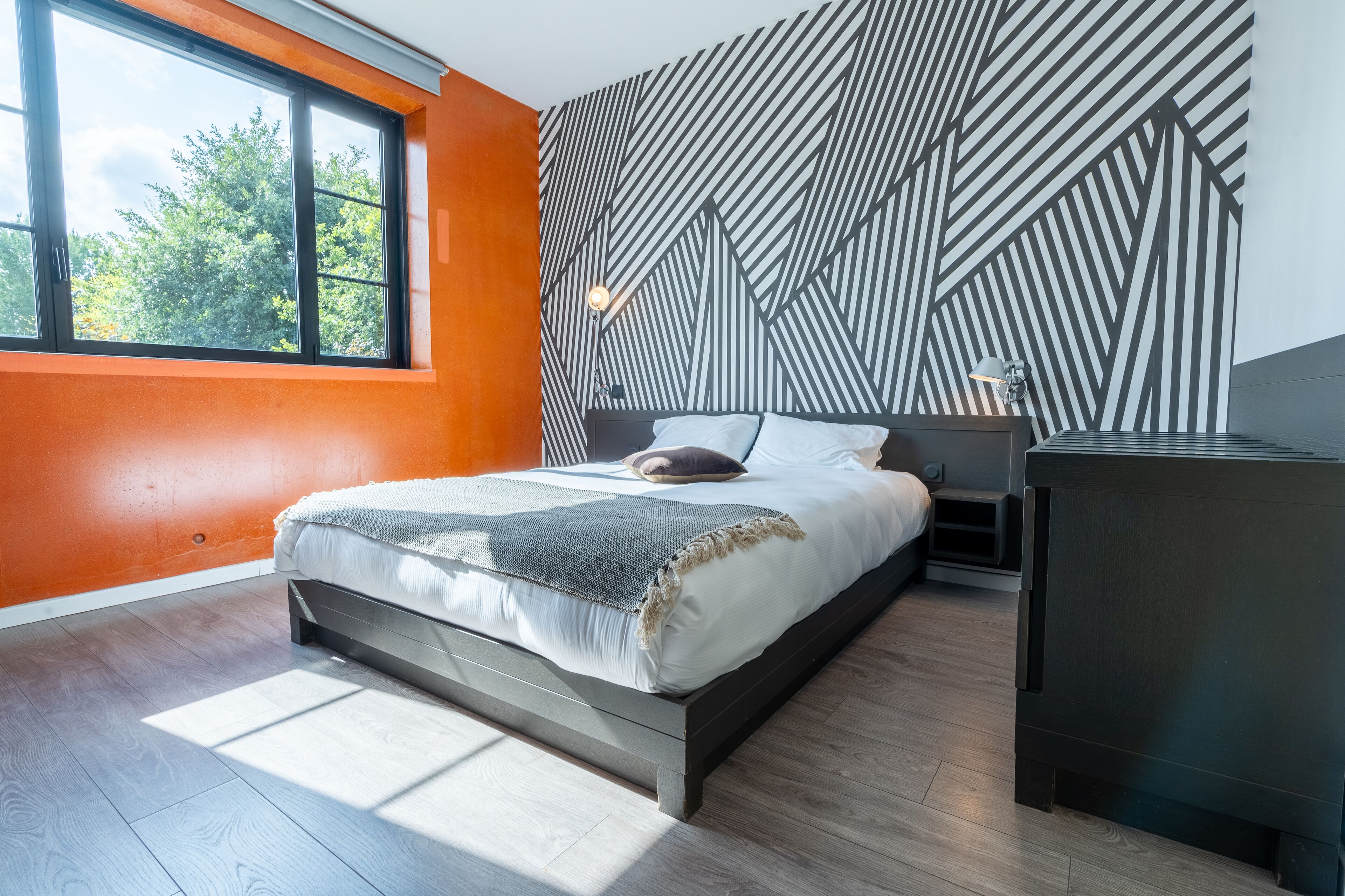 Un dormitorio moderno con un ventanal grande, un muro con estampado geométrico y una cama con cubrecama blanco y gris.