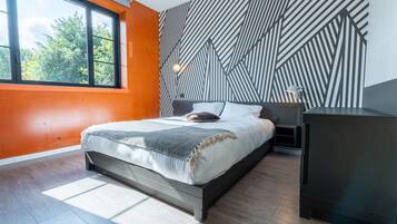 Un dormitorio moderno con un ventanal grande, un muro con estampado geométrico y una cama con cubrecama blanco y gris.