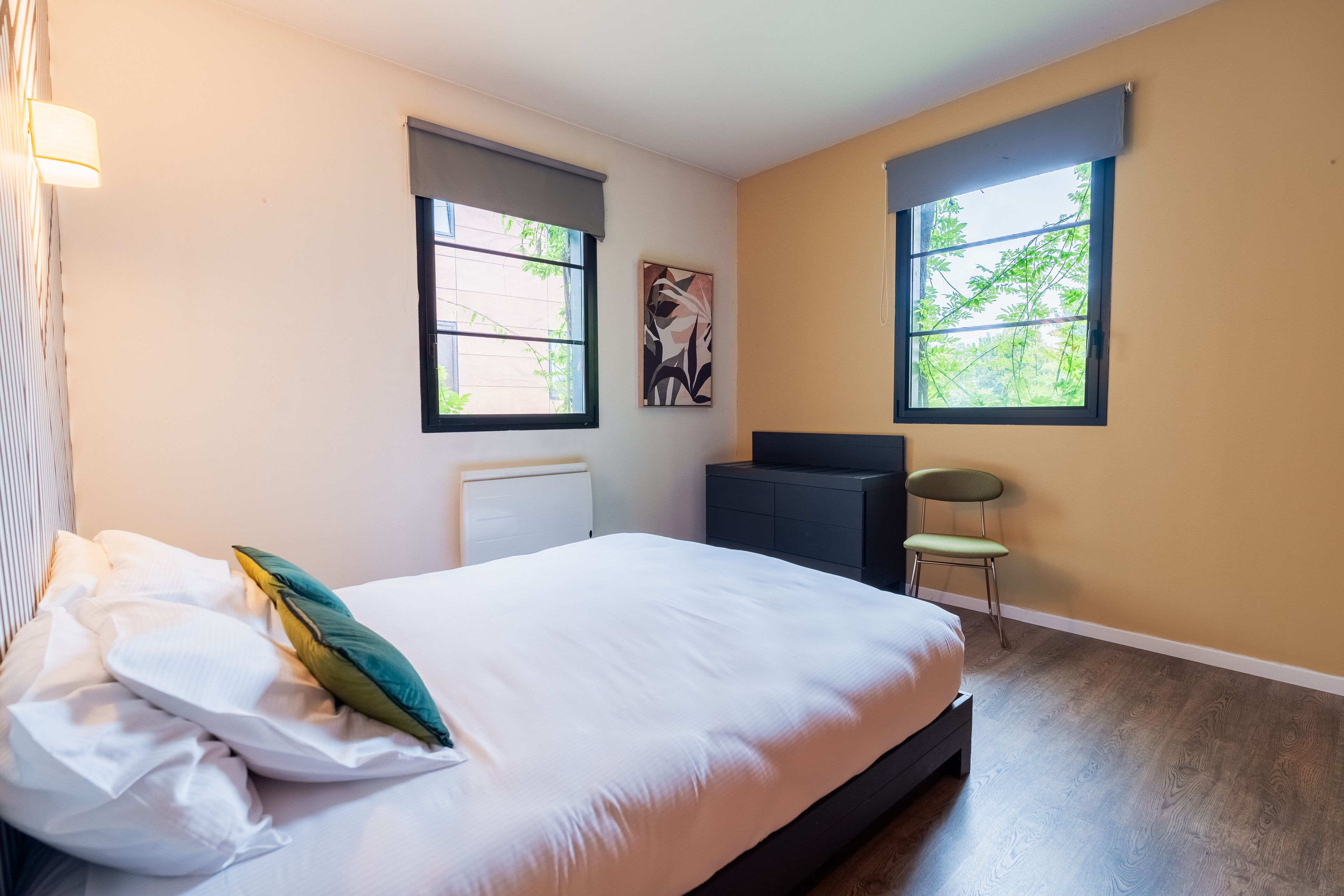 Un dormitorio con cama, dos ventanas, un radiador, una silla y una obra de arte colgada en la pared.