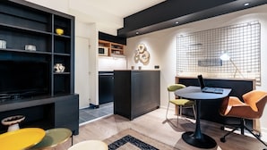 A modern living room with a TV, black shelves, a kitchen area, and a dining table with chairs. - Cowool Lille Villeneuve D'Ascq - Ex Olivarius (Villeneuve-d'Ascq)