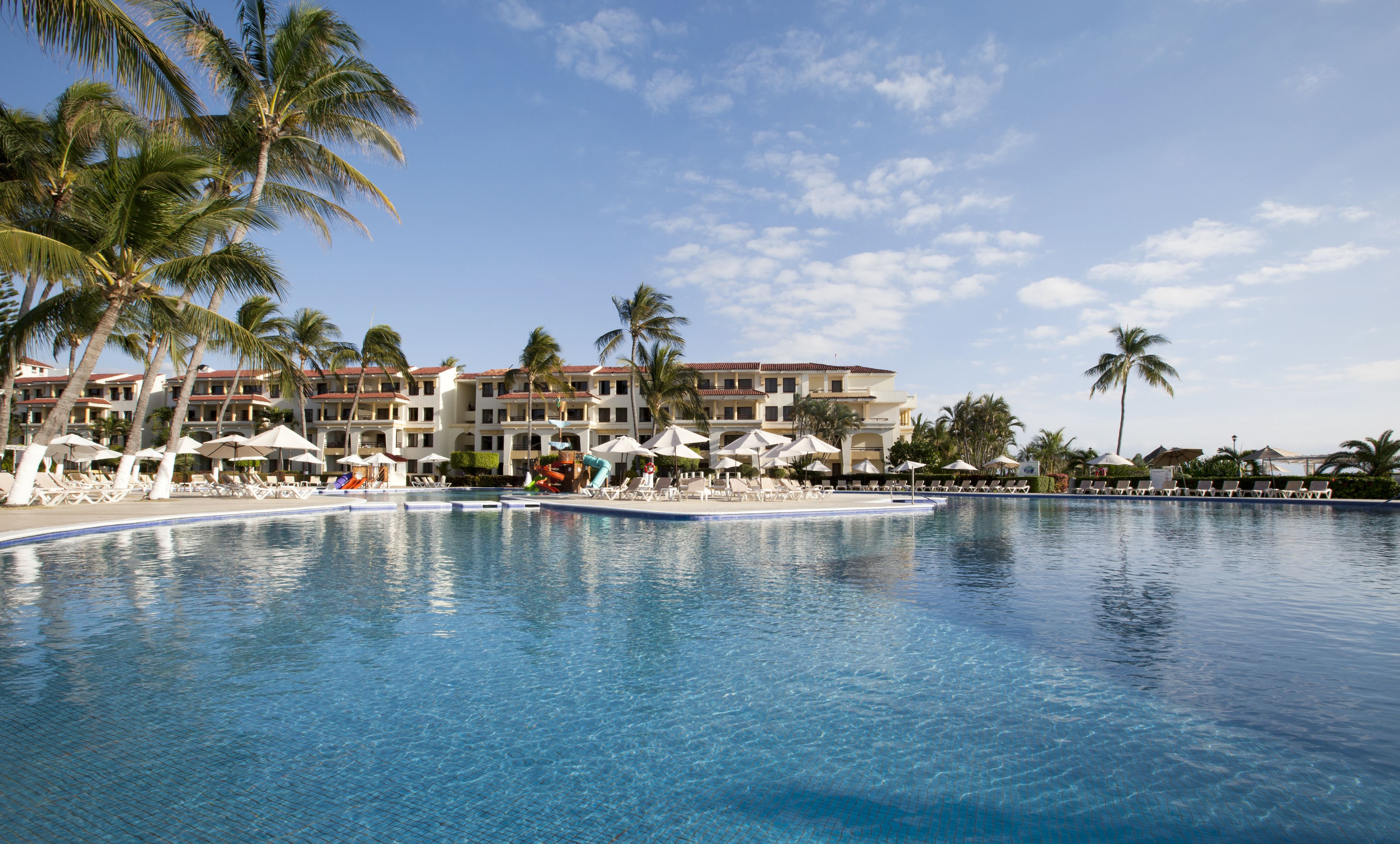 Une piscine de complexe hôtelier, dotée de chaises longues et de parasols, entourée de palmiers et d’un bâtiment d’hôtel.