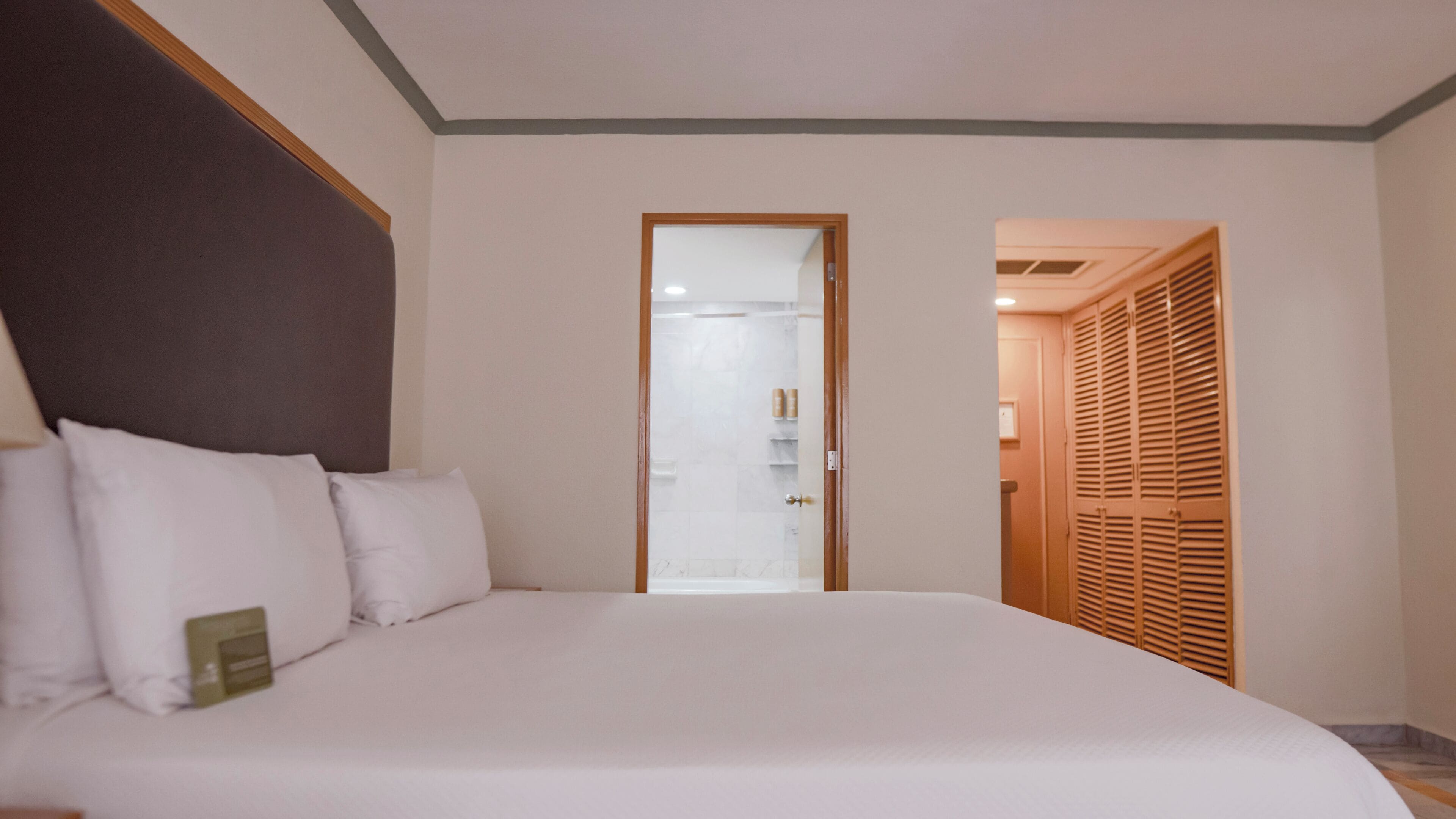 A hotel room with a large bed, a wooden headboard, a bathroom with a glass door, and a wooden wardrobe.