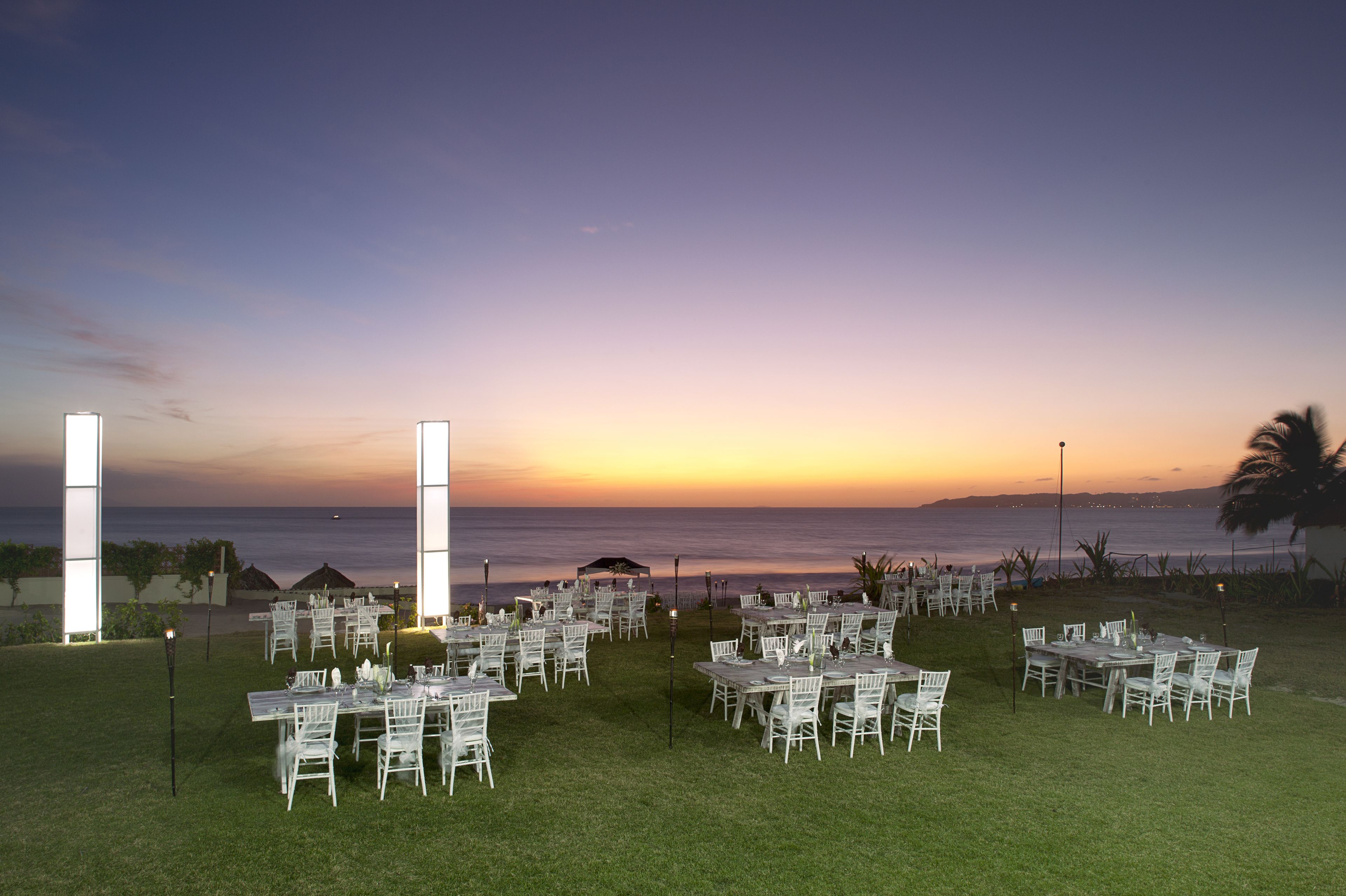 Un espace de restauration en plein air, avec des tables et des chaises disposées sur la pelouse, offrant une vue sur l’océan au coucher du soleil.