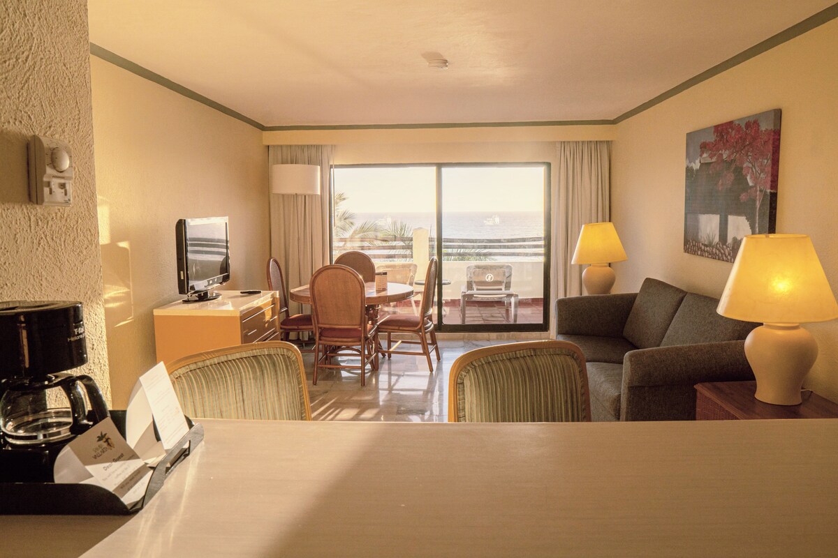 Ein Hotelzimmer mit Essbereich, einer Couch, zwei Lampen, einem Fernseher und Meerblick.