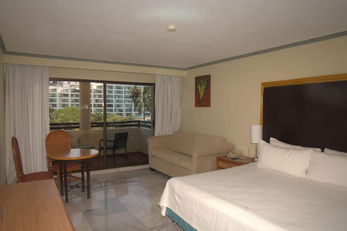 Habitación de hotel con cama, sofá, mesa de comedor con sillas, una mesita con lámpara y vistas al exterior a través de una puerta corredera.
