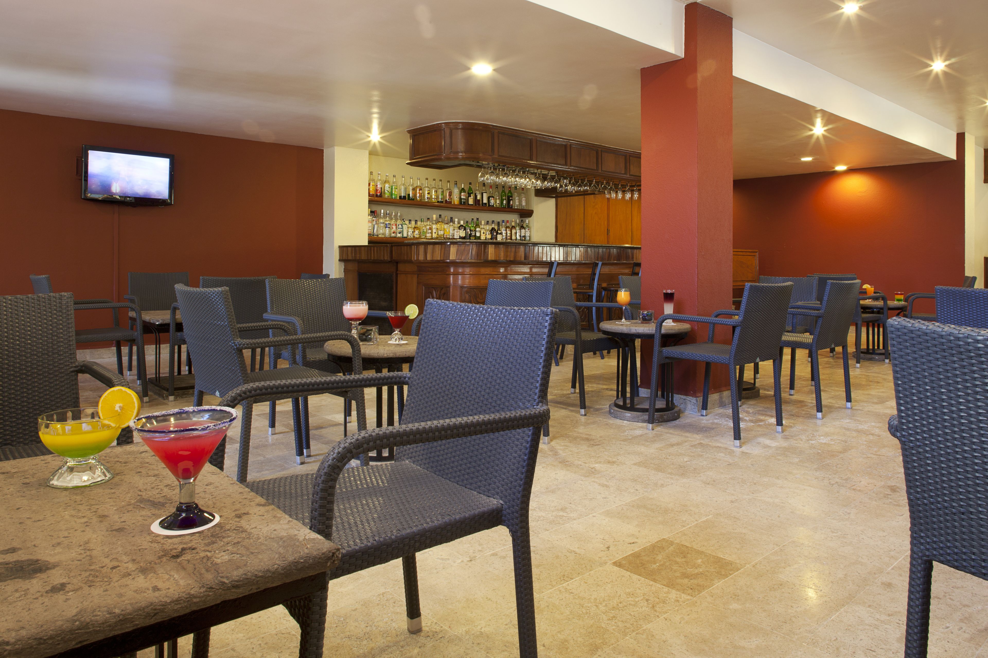 A bar area with multiple seating options, a television mounted on the wall, and a variety of bottles on shelves.