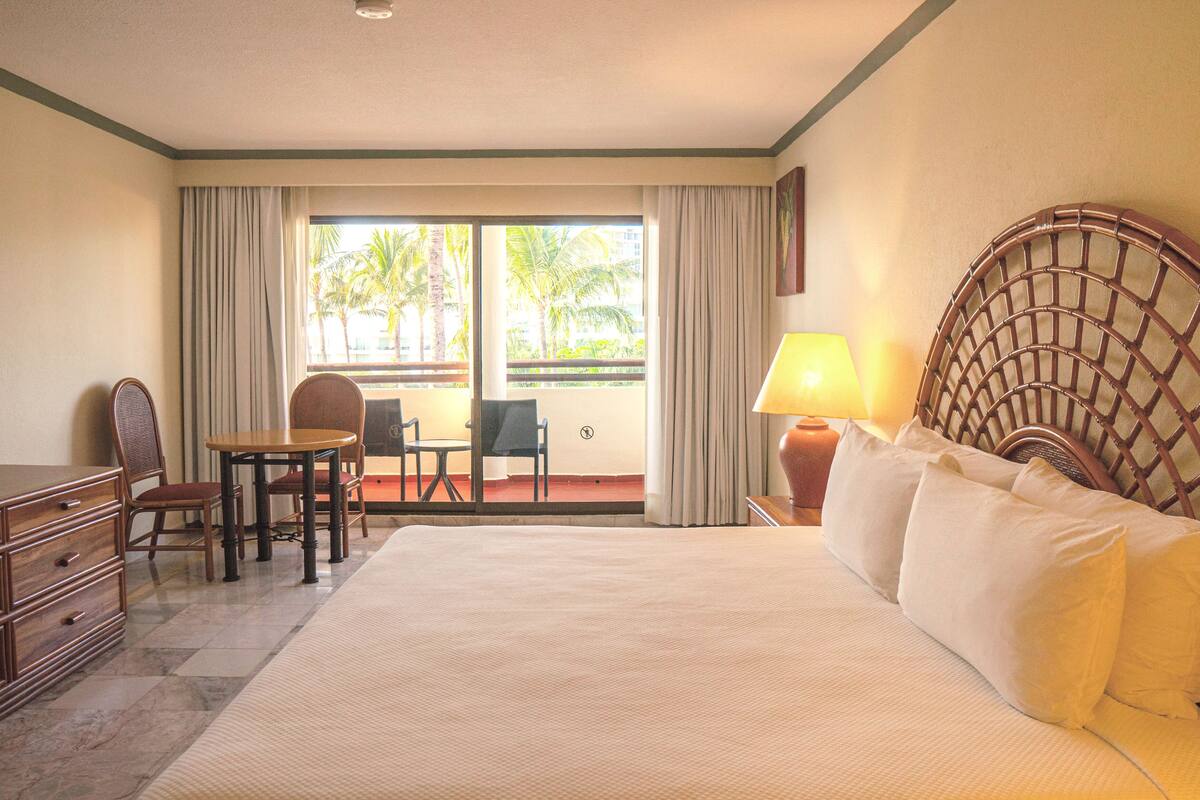 Habitación de hotel con una cama grande, un escritorio con sillas, una lámpara y vistas a palmeras a través del ventanal.