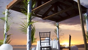 A beachside gazebo with a table and chairs, surrounded by palm trees and white fabric decorations. - Samba Vallarta - All Inclusive (Nuevo Nayarit)