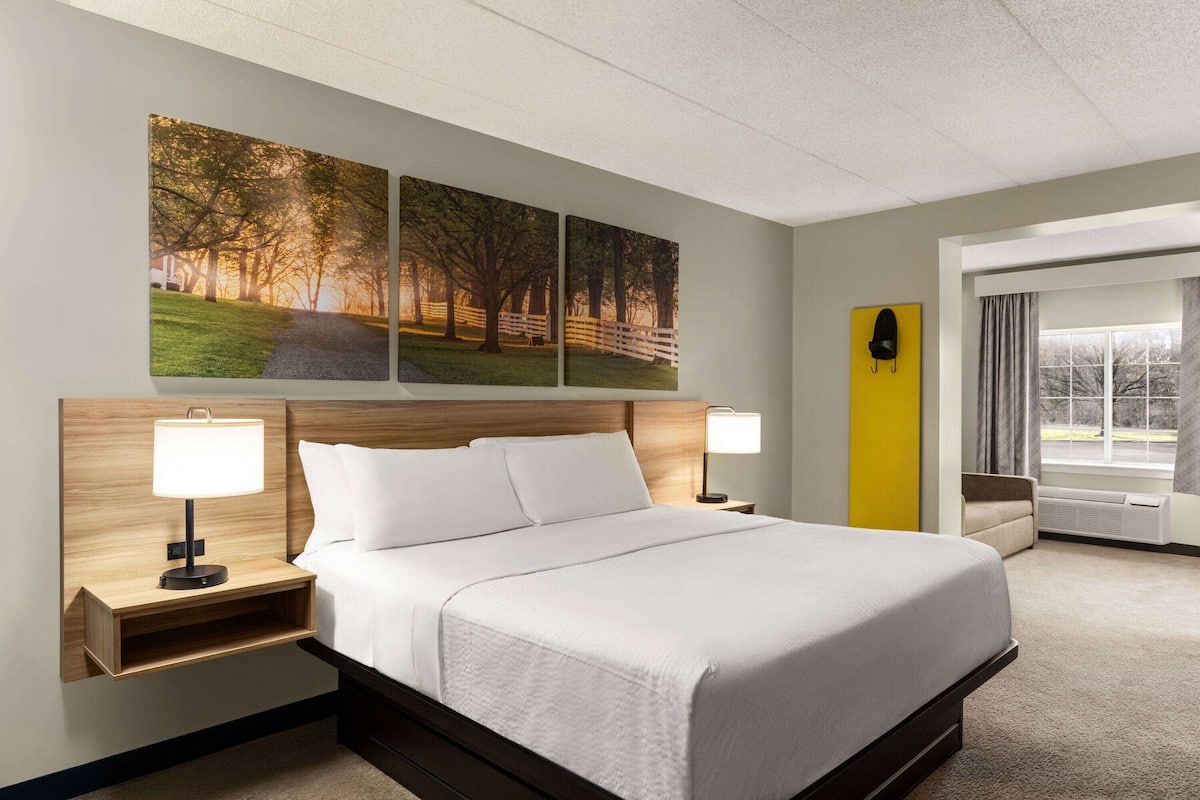 Ein Hotelzimmer mit einem großen Bett, zwei Nachttischen mit Lampen, einer Kopfteil, einem Fenster mit Vorhängen und drei Wandbildern.