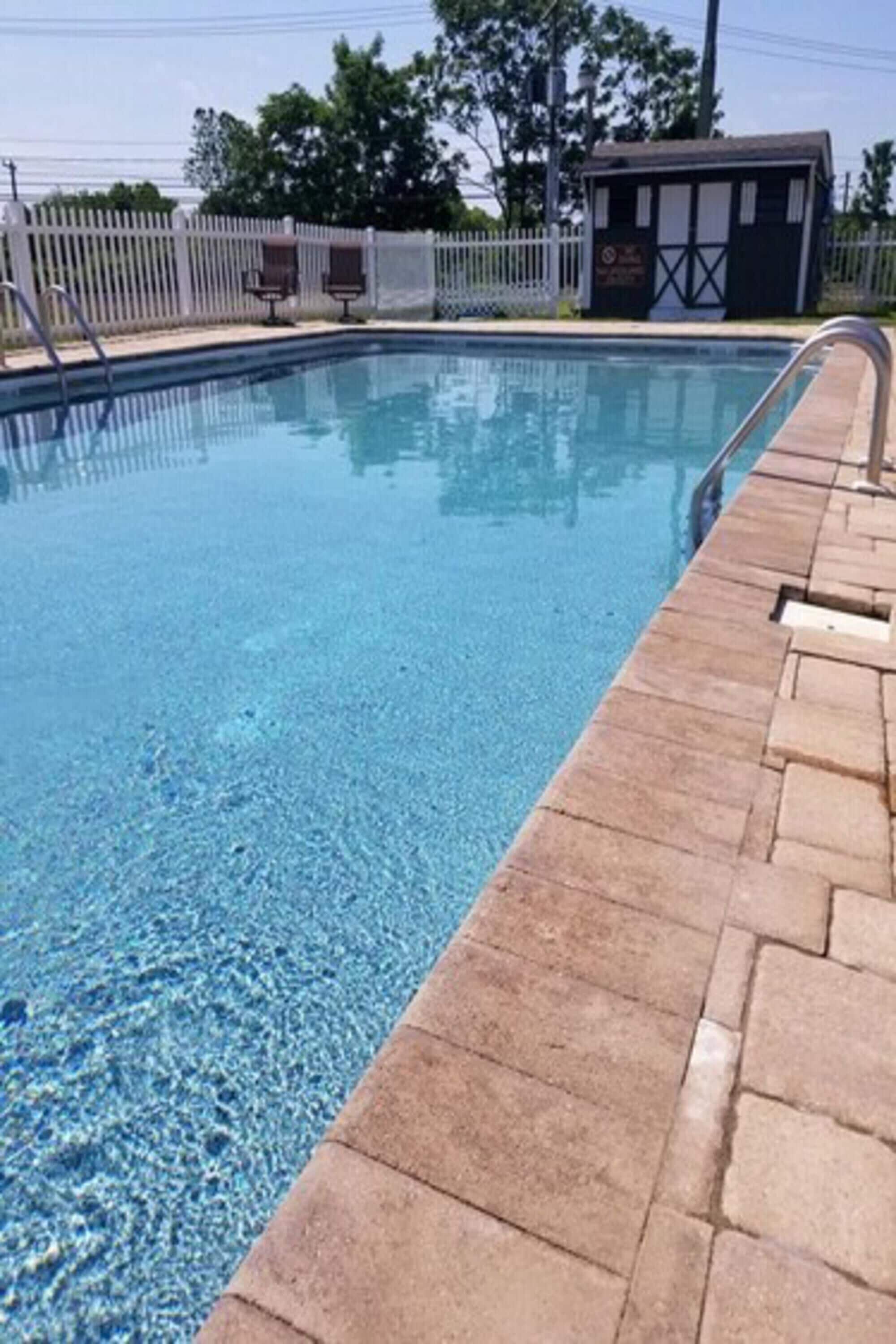A clear swimming pool with a stone border, a small building in the background, and a white fence.
