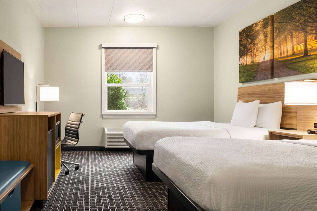 Ein Hotelzimmer mit zwei Betten, einem Schreibtisch, einem Stuhl, einem Fernseher und einem Fenster mit Blick auf Bäume.