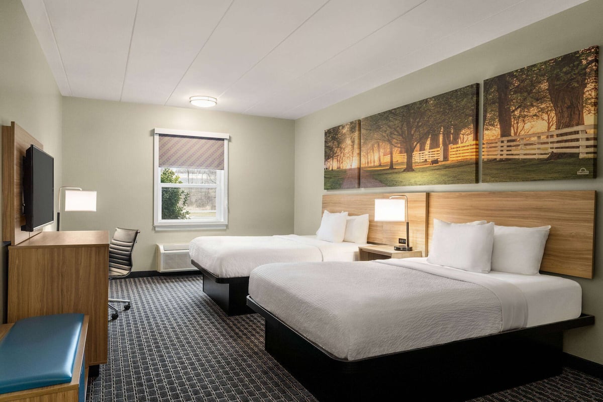 Ein Hotelzimmer mit zwei Betten, einem Schreibtisch, einem Stuhl, einem Fernseher und einem Fenster mit Jalousien.