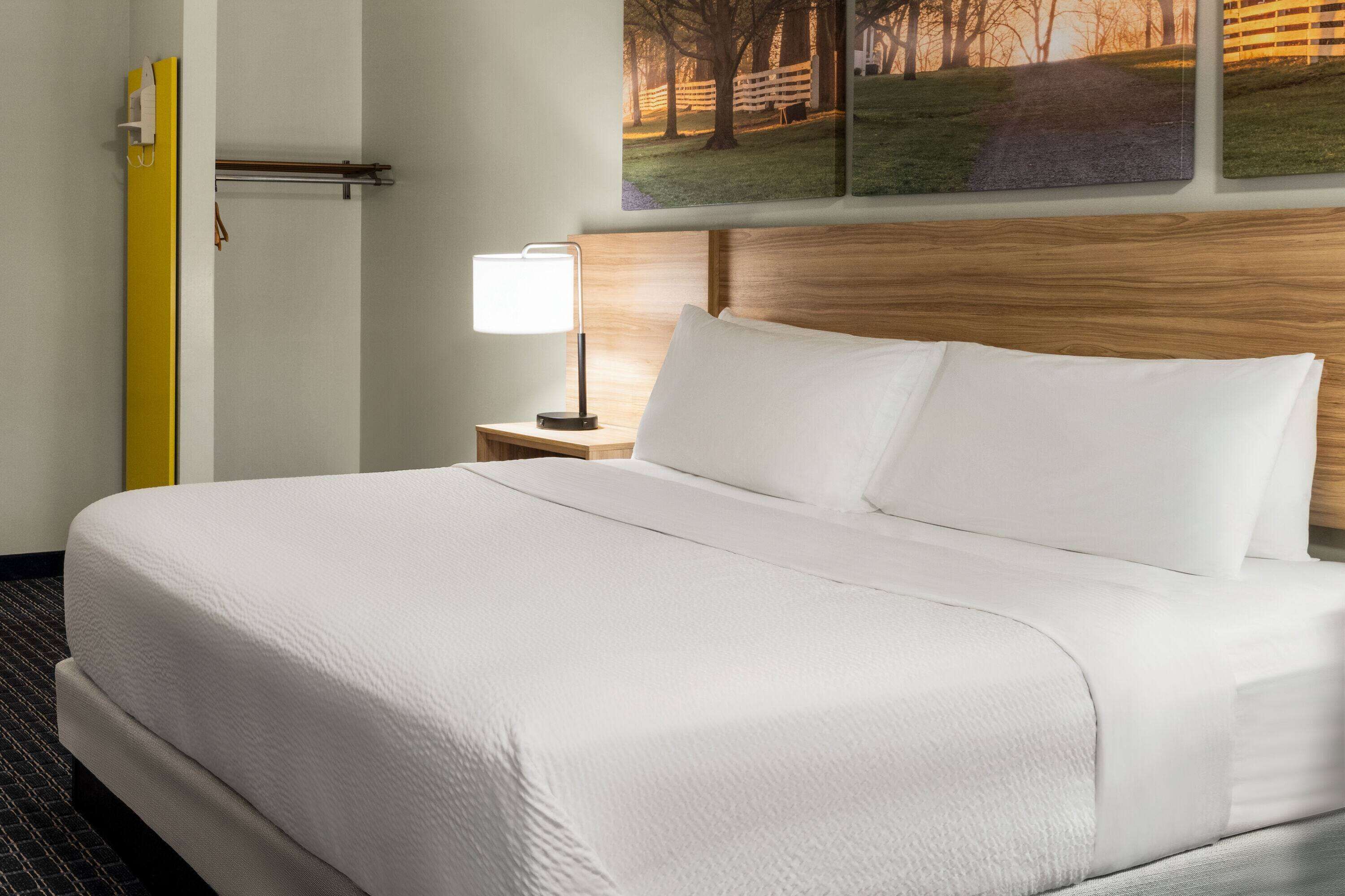 Una cama bien hecha con sábanas blancas, un cabecero de madera y una mesita de noche con una lámpara.