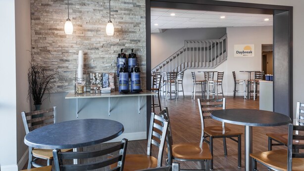Un café moderno con pared de ladrillo, mesas y sillas redondas. Se observan dispensadores de café y un letrero que anuncia 'Daybreak Breakfast'.