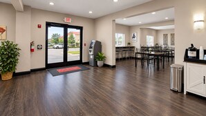 A hotel lobby with a reception desk, a self-service kiosk, a dining area with tables and chairs, and a trash can.