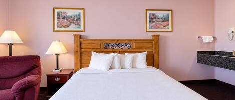 Habitación de hotel con una cama, dos lámparas de noche, un cabecero de madera, un sillón rojo y dos cuadros en la pared.