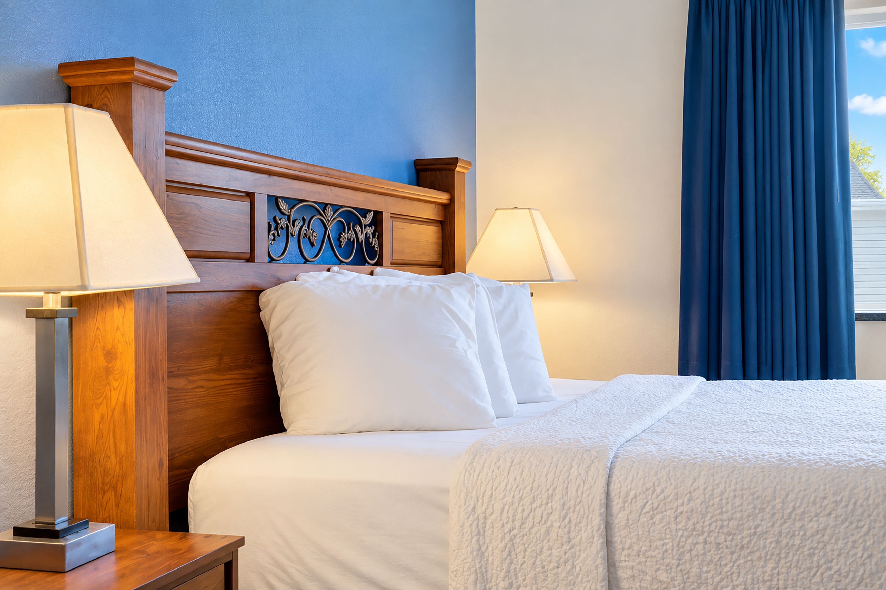 Quarto de hotel com cabeceira de madeira, duas luminárias de apoio, cama com roupa de cama branca e janela com cortinas azuis.