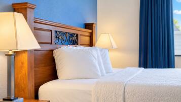 Quarto de hotel com cabeceira de madeira, duas luminárias de apoio, cama com roupa de cama branca e janela com cortinas azuis.