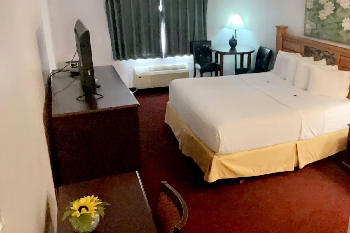 Una habitación de hotel con una cama, un escritorio con computadora, una silla, una lámpara y un cuadro en la pared.