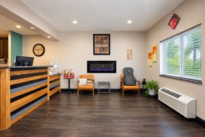 A reception area with a reception desk, a clock, a television, two chairs, a fan, and a window with blinds. - Fairbridge Inn & Suites Williston Near Airport (Williston)