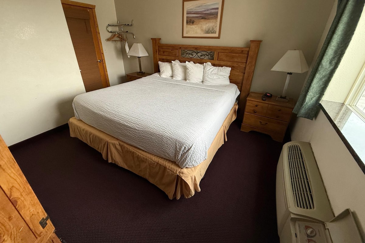 A single bed with a white comforter and a wooden headboard, a bedside table with a lamp, a wall-mounted heater, and a door to the left.