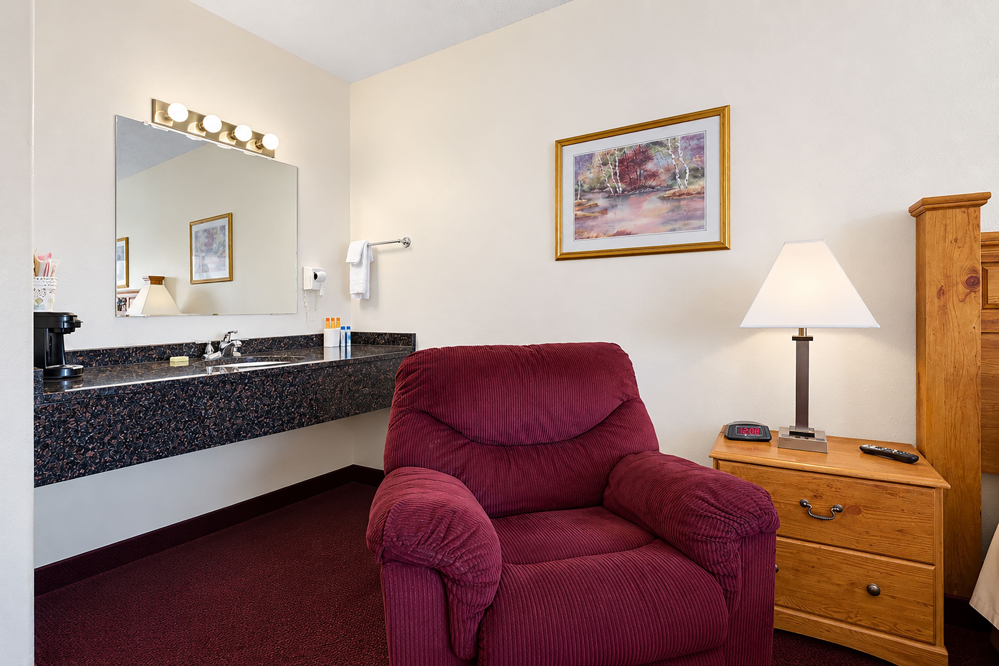 A hotel room with a marble bathroom vanity, a red upholstered chair, a wooden nightstand with a lamp, and a painting on the wall.