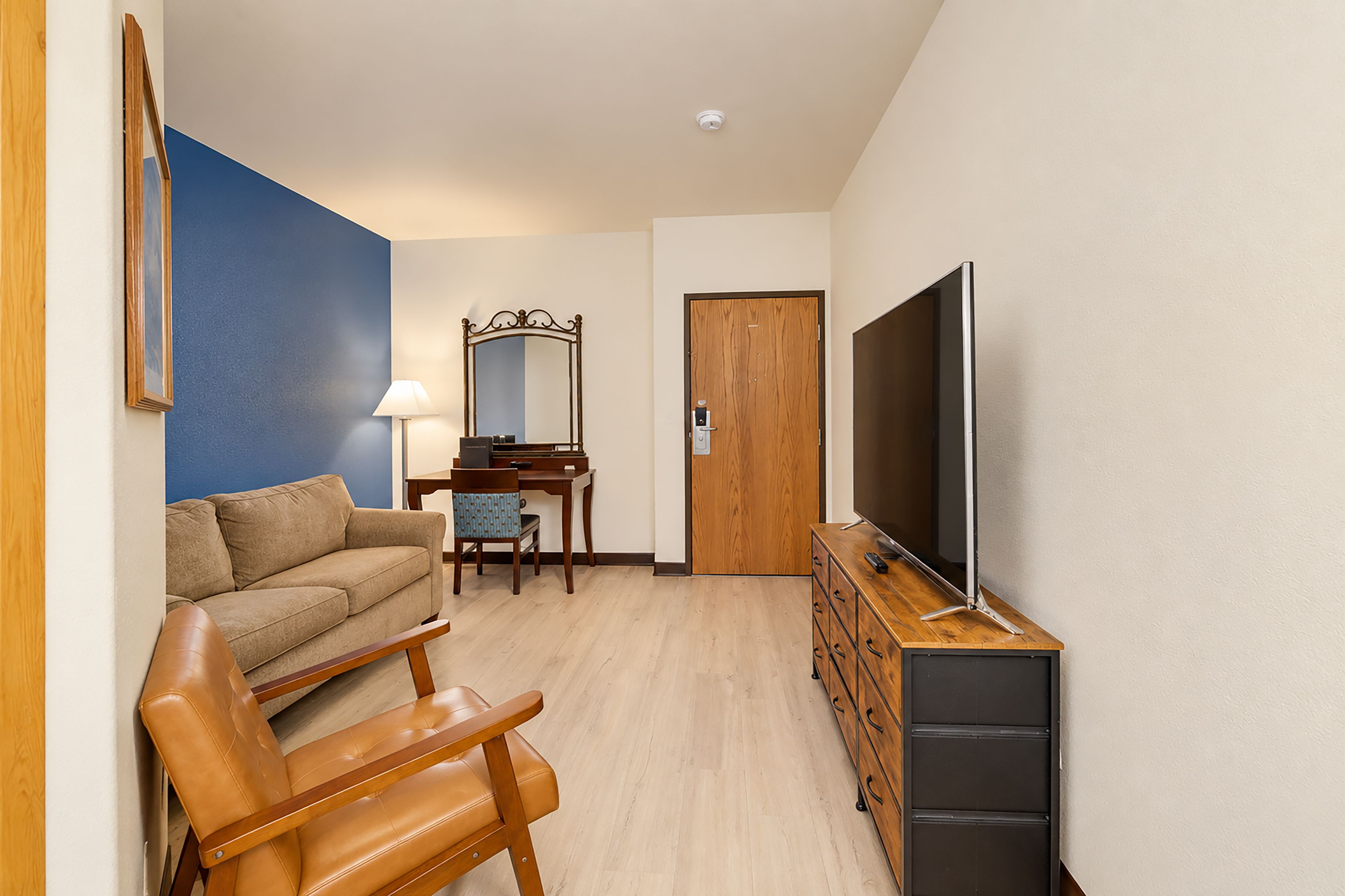 Una sala de estar moderna con un sofá, una silla de madera, un escritorio con una lámpara, un televisor sobre un mueble de madera y una puerta.