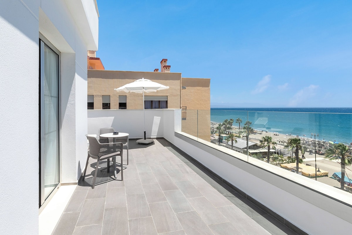 A balcony with a table and chairs, a white umbrella, and a view of the beach and ocean.