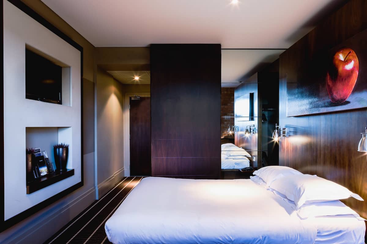 A modern hotel room with a large bed, a television mounted on the wall, a painting of an apple, and a nightstand with a lamp.