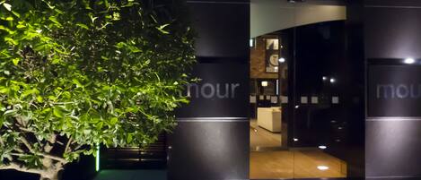 Entrada moderna de un edificio con puertas de cristal, un árbol a la izquierda y luz interior visible a través de las puertas.