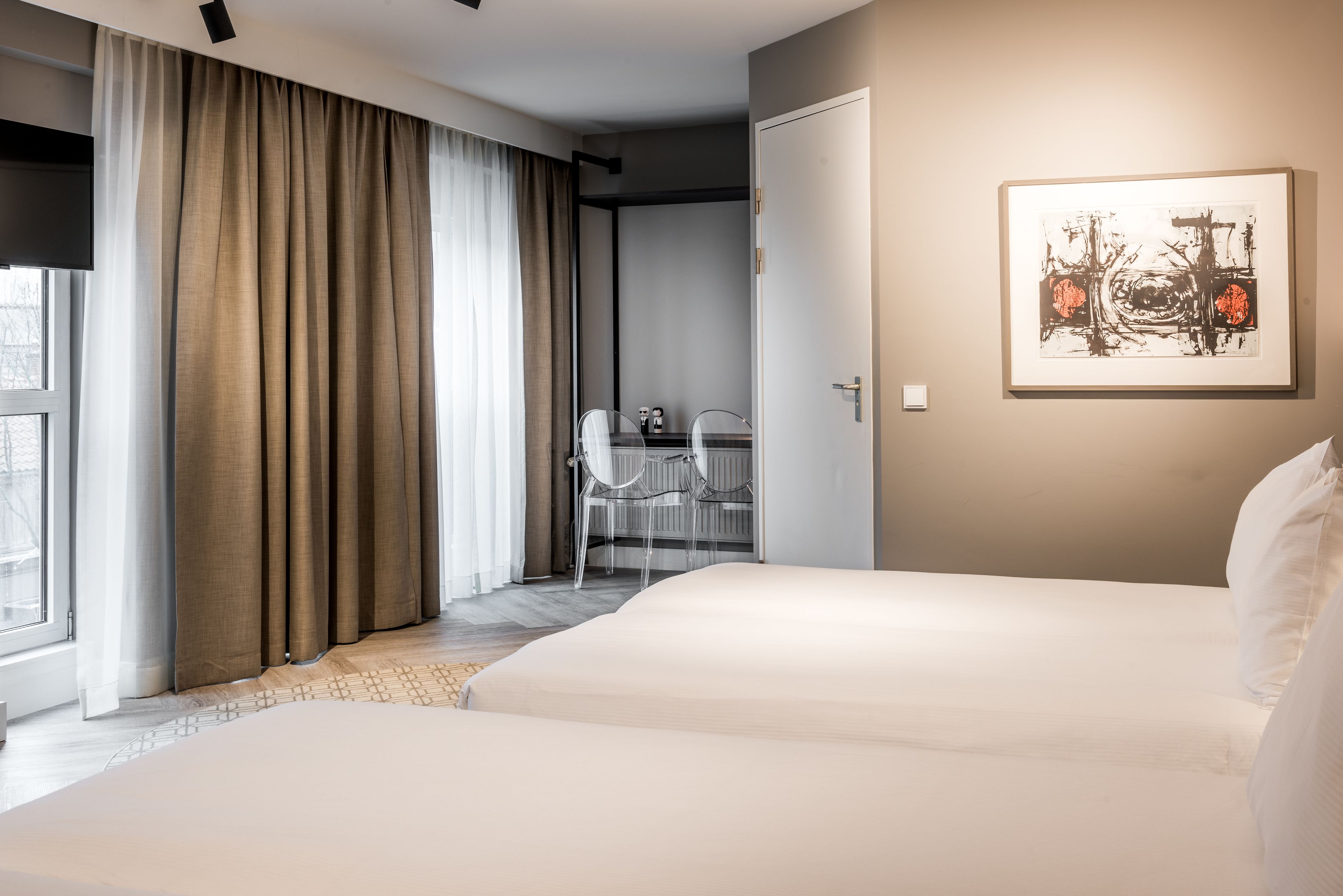 A hotel room with two beds, a desk with a chair, a window with curtains, and a framed artwork on the wall.