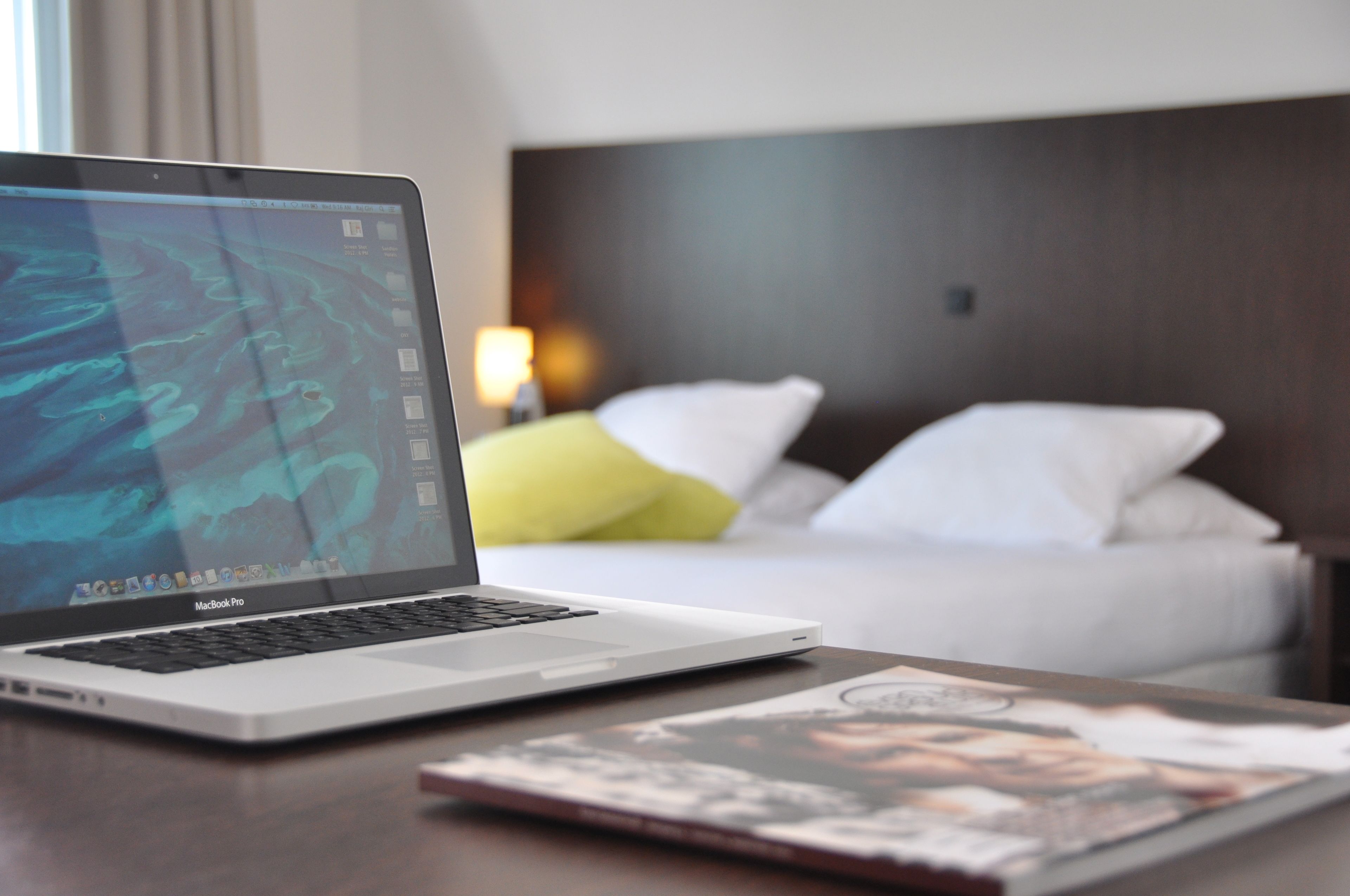Um MacBook Pro sobre uma mesa de madeira, uma revista, uma cama com almofadas brancas e amarelas e uma mesa de cabeceira com uma luminária em um quarto de hotel.