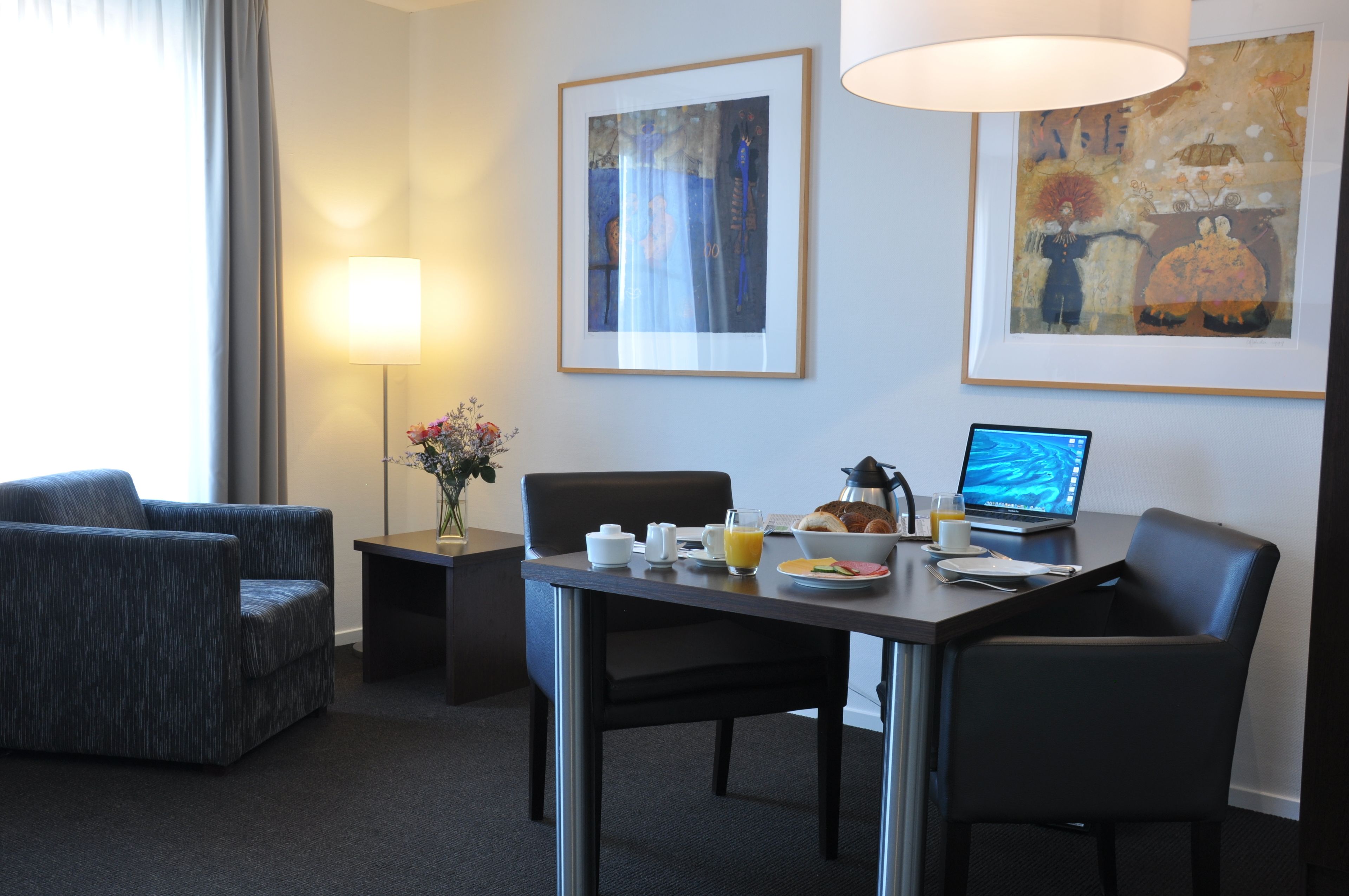 A hotel room with a dining table, chairs, a laptop, a lamp, and artwork on the walls.