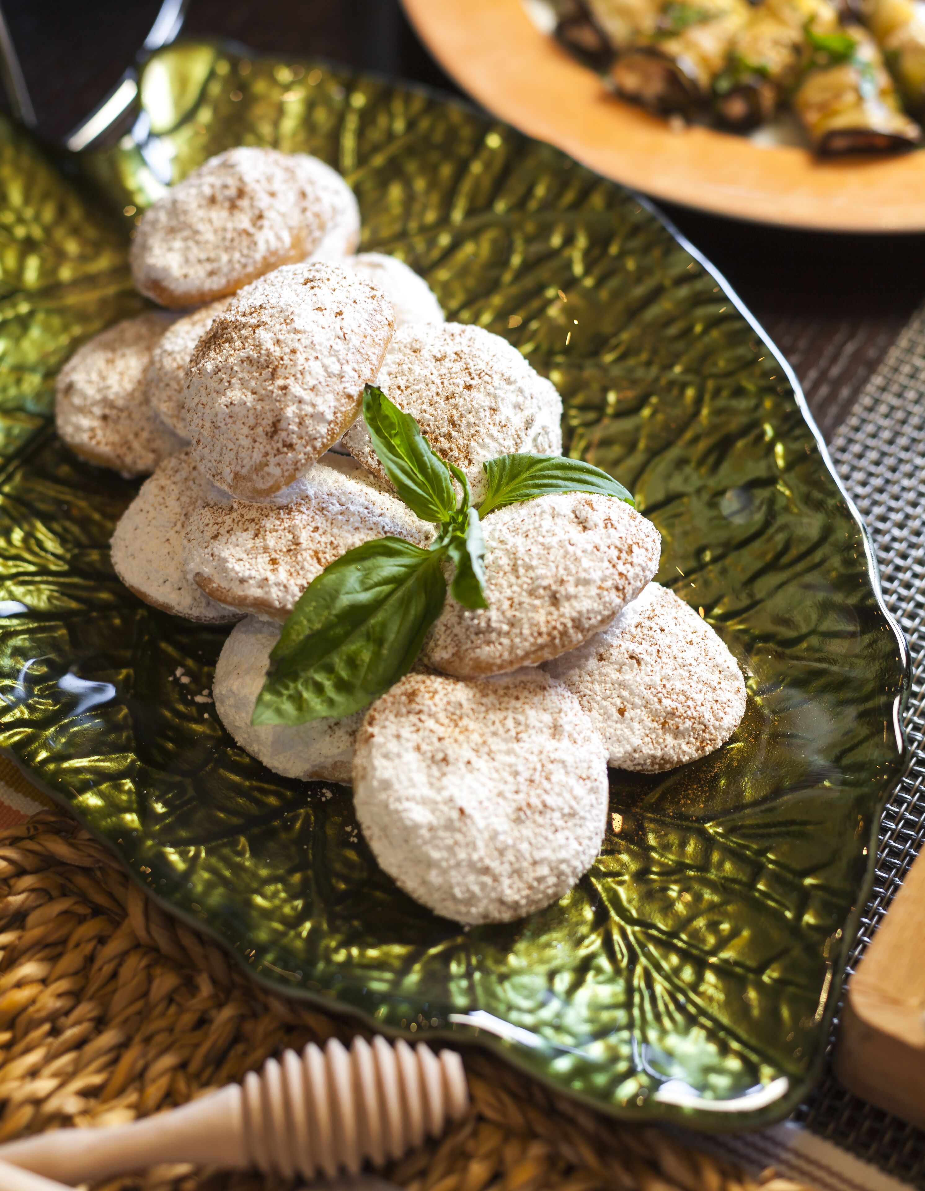 A plate of round cookies dusted with powdered sugar and a sprig of basil, placed on a green glass plate.