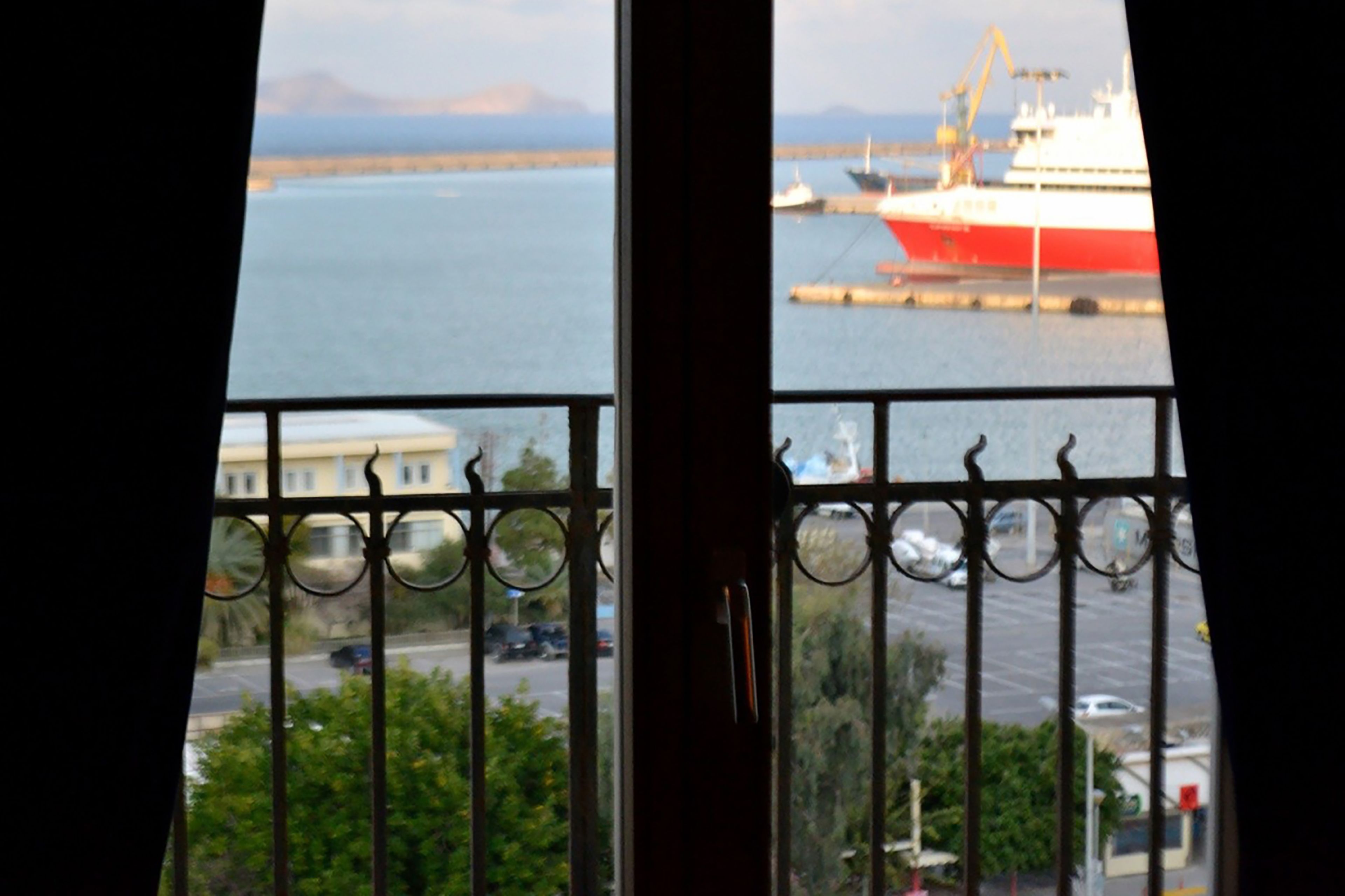 A view through a window with a metal railing, featuring a large ship docked at a port, a road with cars, and some greenery in the foreground.