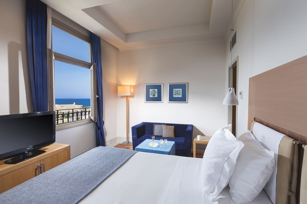 A hotel room with a bed, a TV, a desk, a chair, a lamp, and a view of the sea.