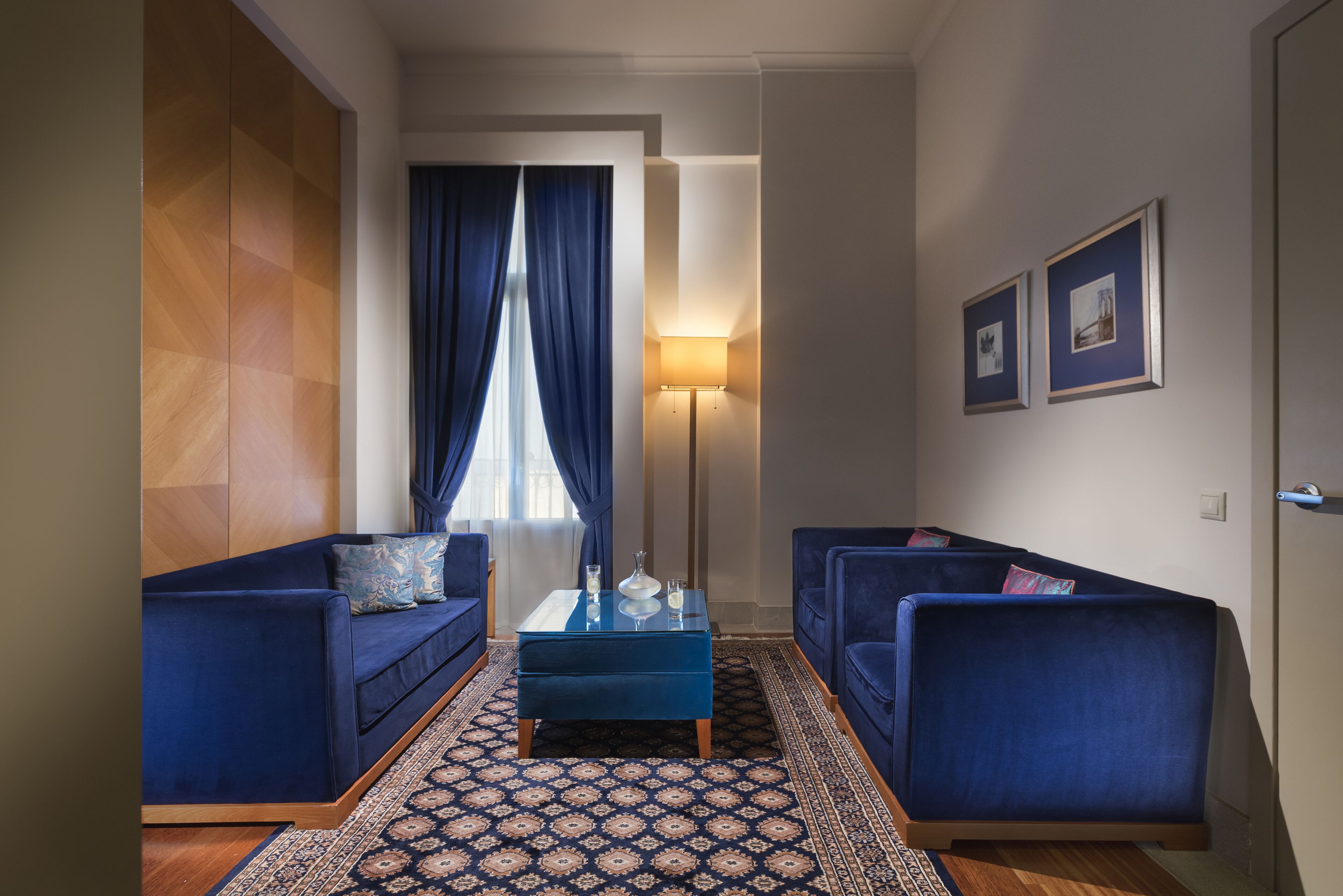 A modern living room with two blue sofas, a matching ottoman, a patterned rug, a standing lamp, and framed pictures on the wall.