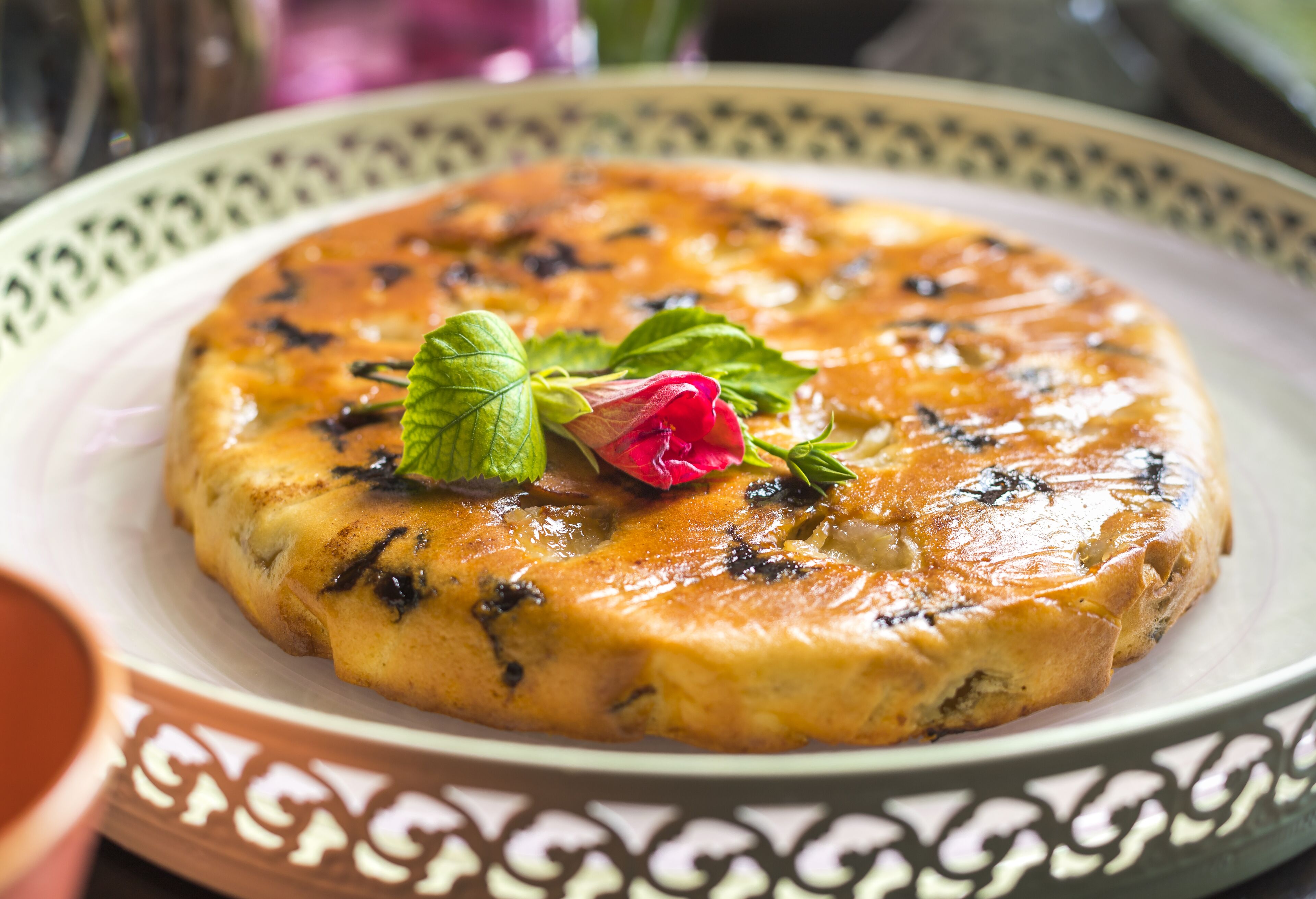 A round dish with a golden-brown crust, garnished with a mint leaf and a pink flower, on a decorative plate.