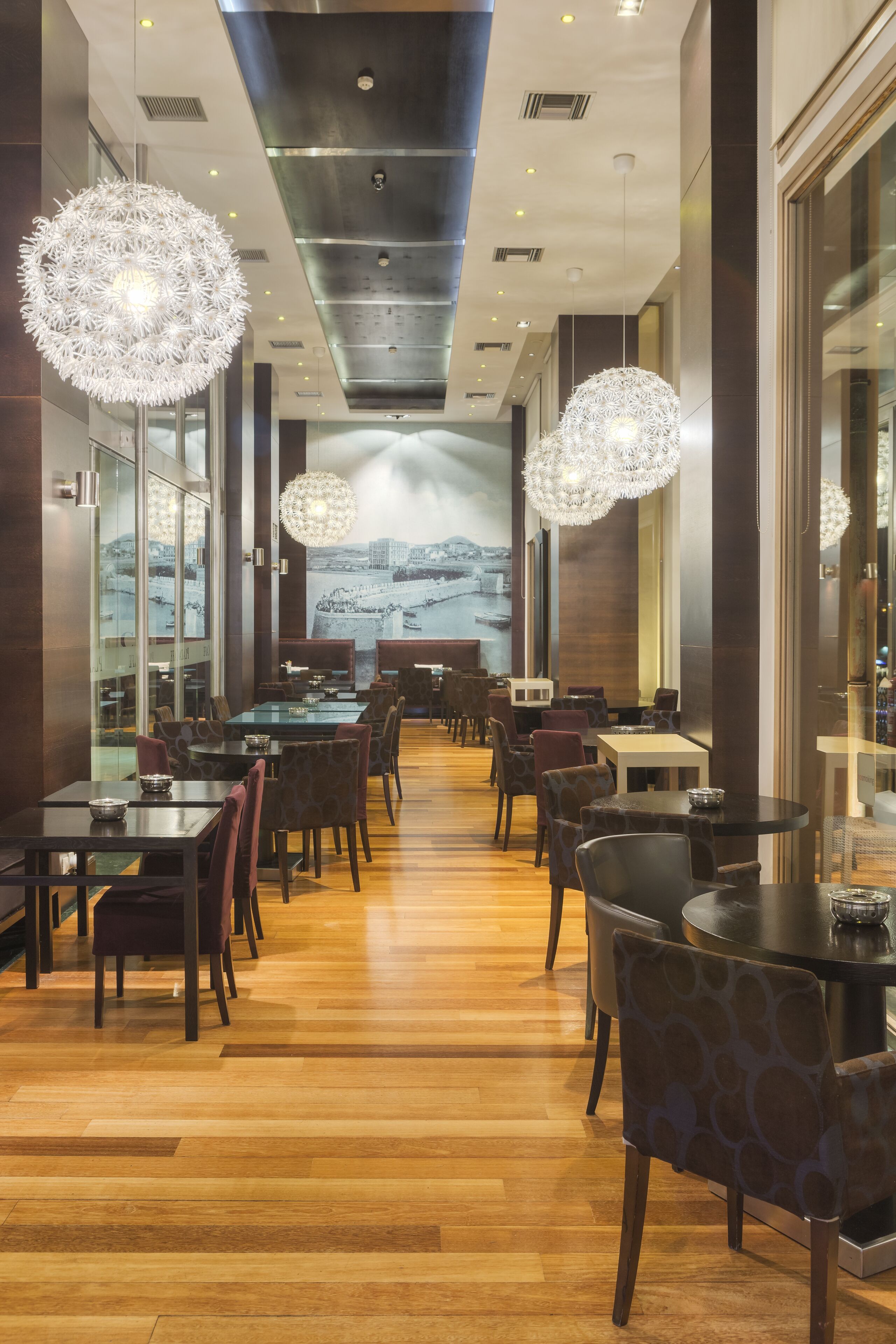 A modern restaurant with a long wooden floor, round tables, and chairs. There are large chandeliers hanging from the ceiling.