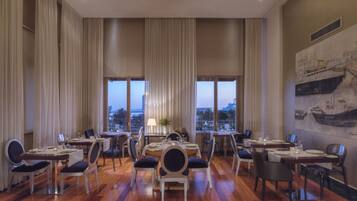 Un comedor con varias mesas y sillas, ventanas amplias con cortinas y vistas a la ciudad.