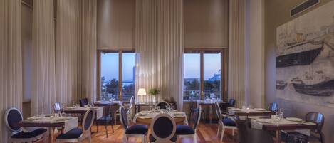 Un comedor con varias mesas y sillas, ventanas amplias con cortinas y vistas a la ciudad.