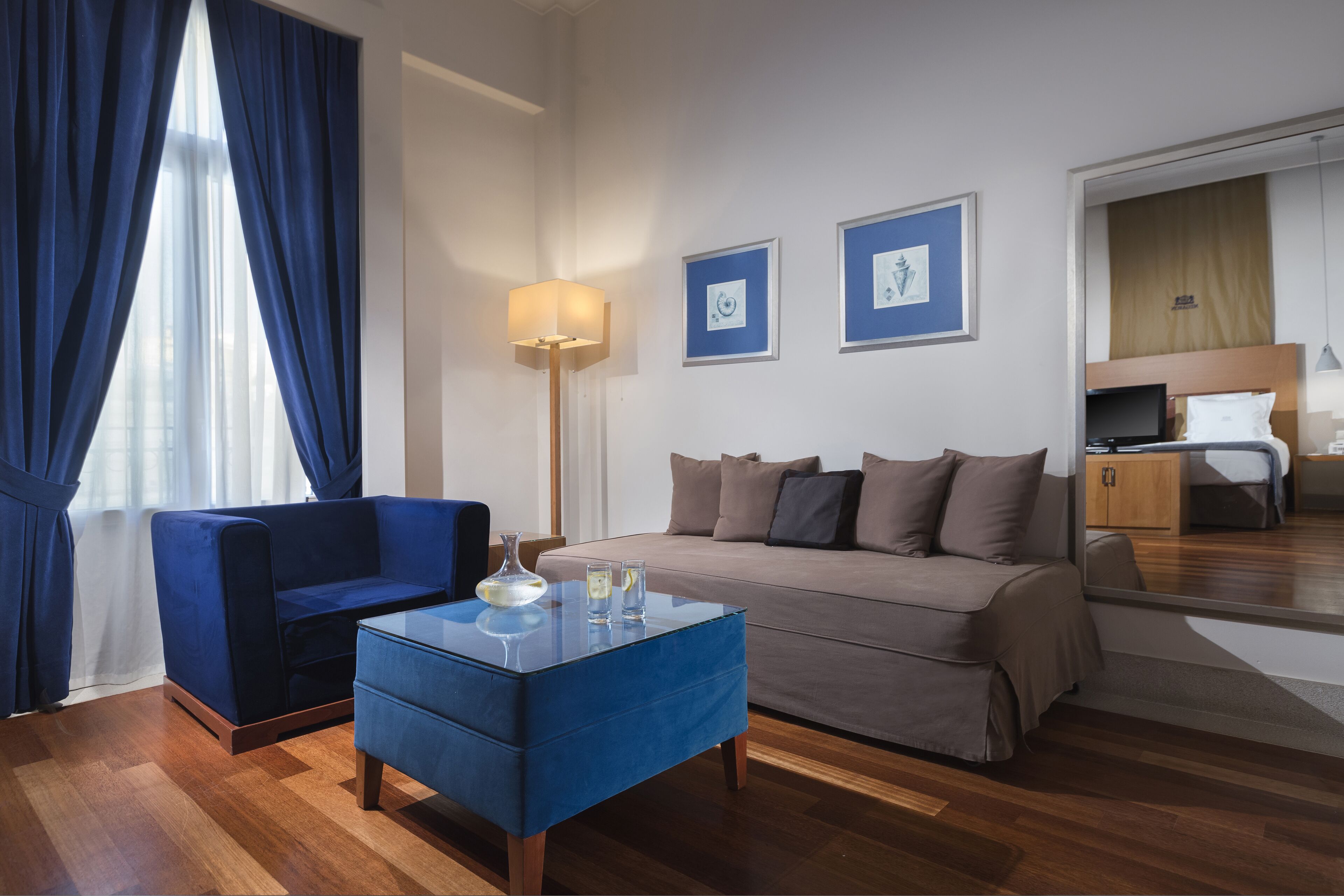 A modern hotel room with a blue sofa, a glass coffee table, a beige sofa, a floor lamp, and two framed pictures on the wall.