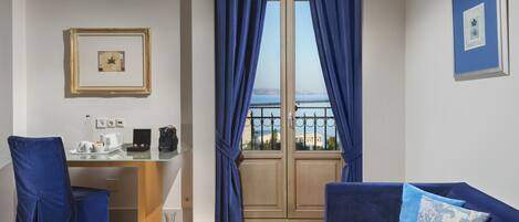 A room with a desk, a chair, a sofa, a view of the sea, and a framed picture on the wall.
