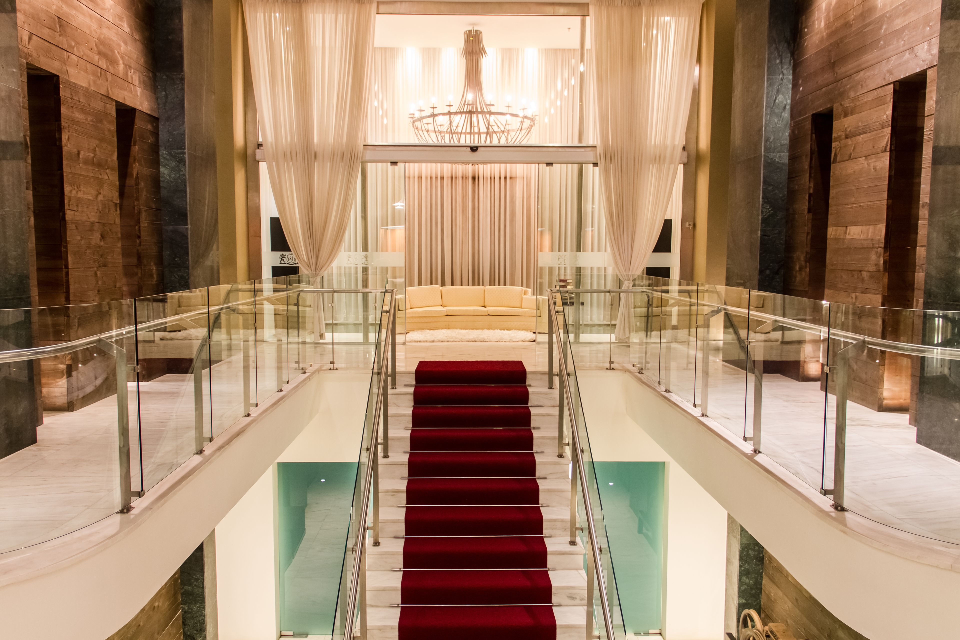 Una gran escalera con un sendero de alfombra roja que conduce a un sofá lujoso, rodeado de barandas de cristal y cortinas colgantes.