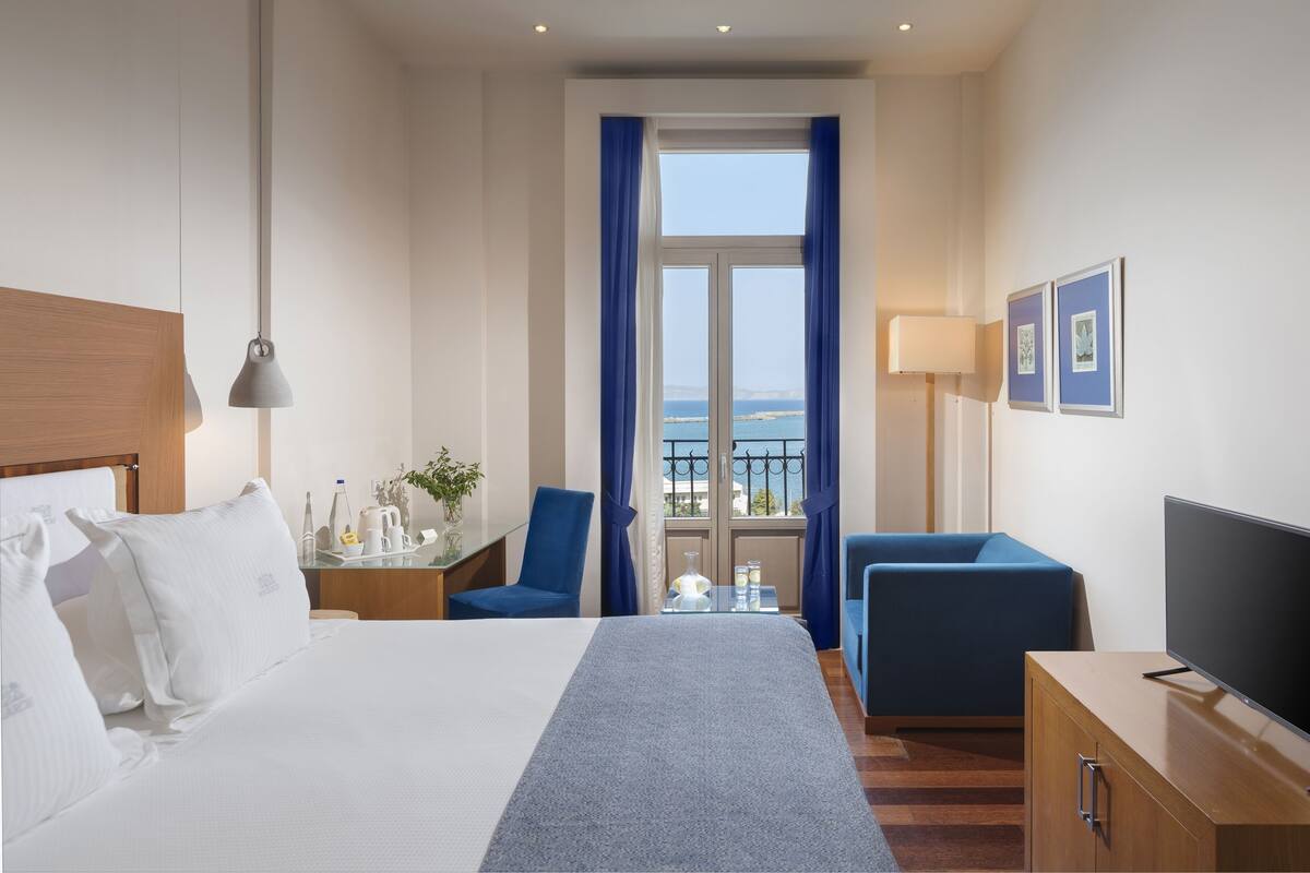 A hotel room with a bed, a desk with a chair, a TV, and a view of the sea through a window.