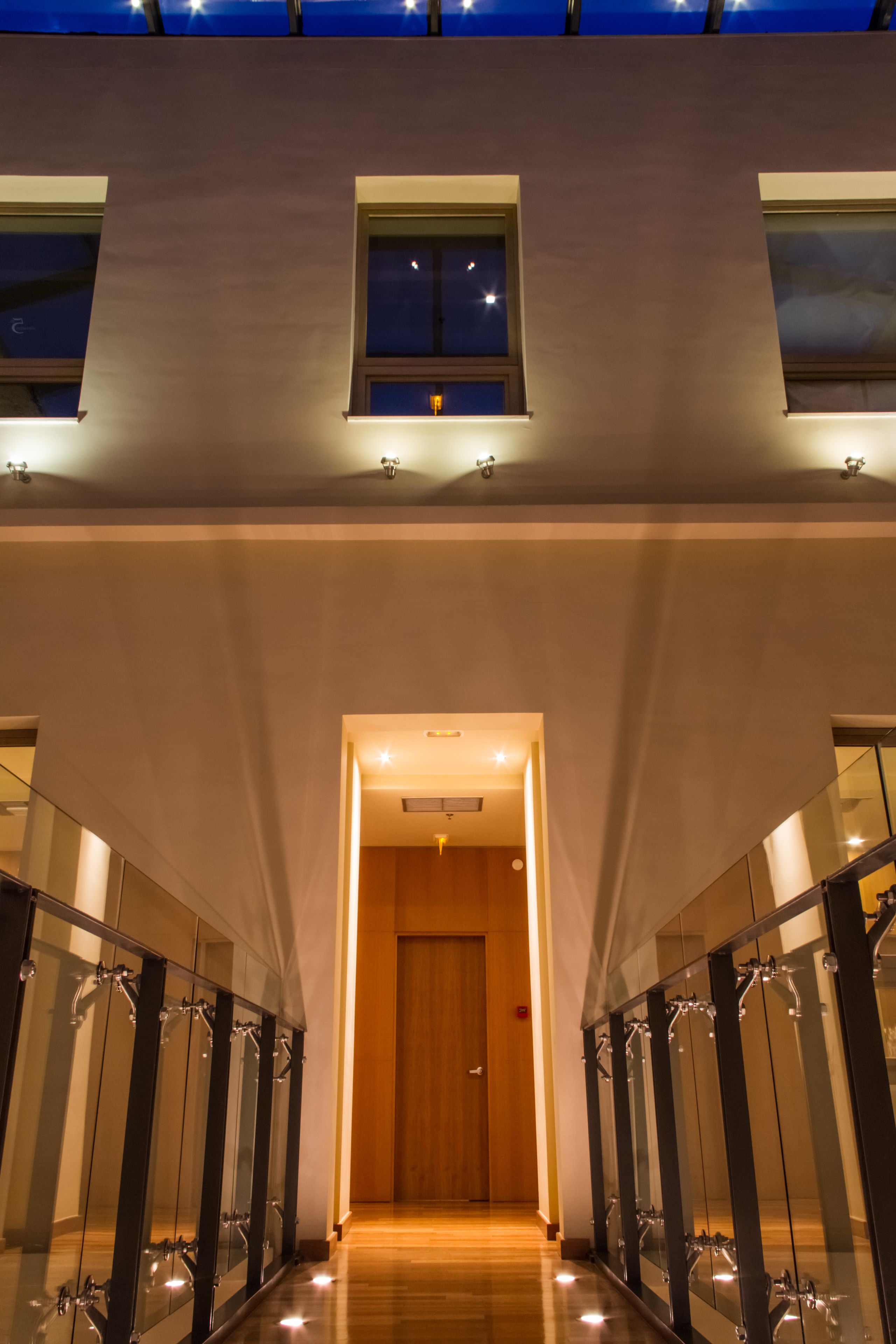 Un couloir d’hôtel moderne avec des rampes de protection en verre, une porte en bois au fond et une lucarne au plafond.