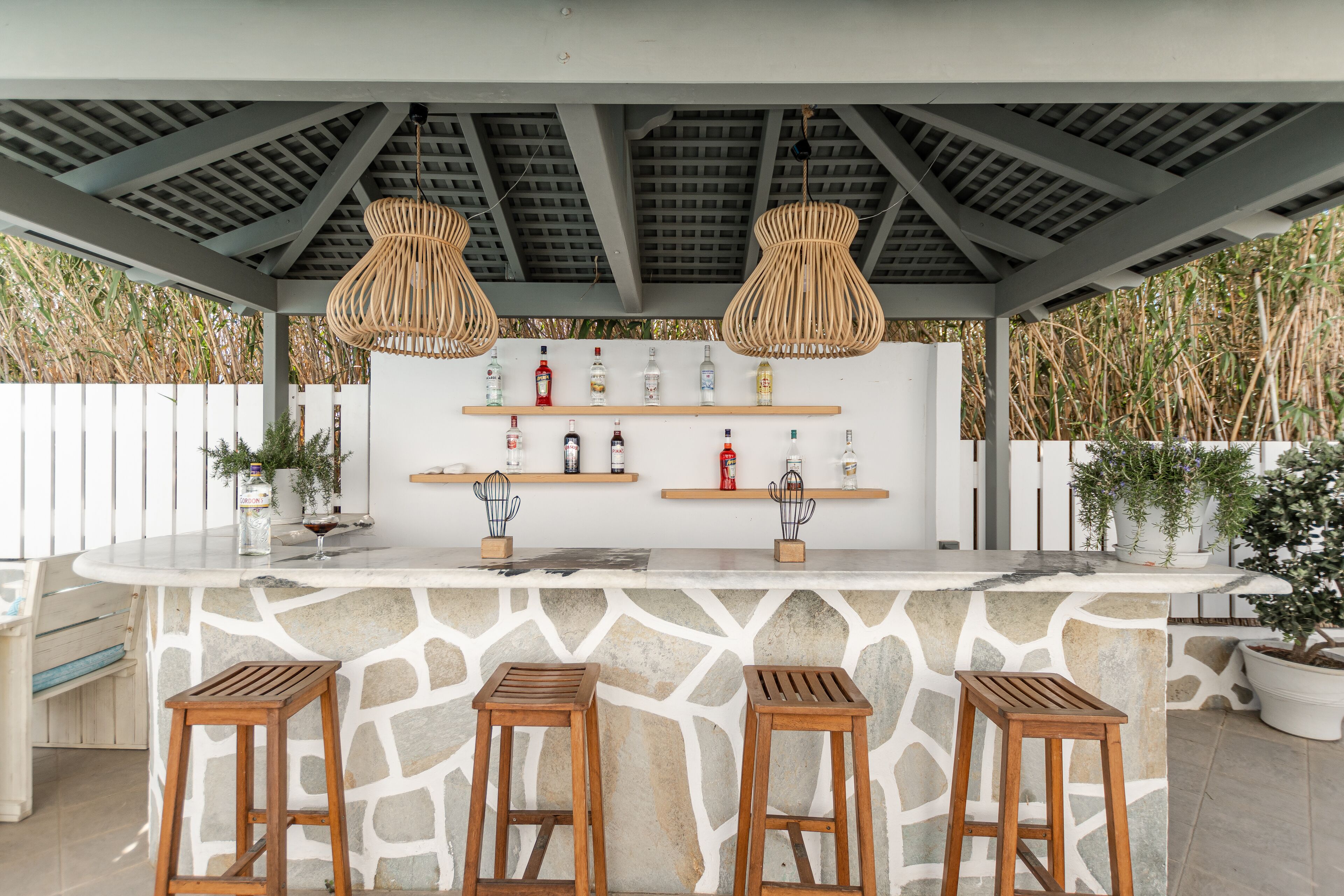 A covered outdoor bar area with a stone counter, wooden stools, hanging light fixtures, and shelves with bottles and plants.