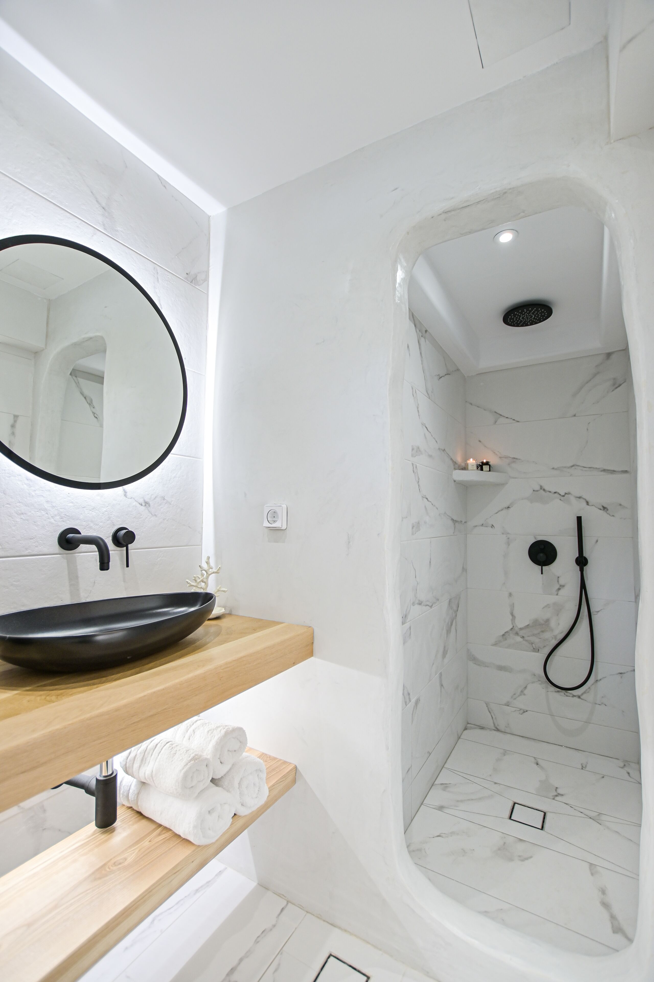 A modern bathroom with a round mirror, black sink, wooden countertop, and a walk-in shower with marble tiles.