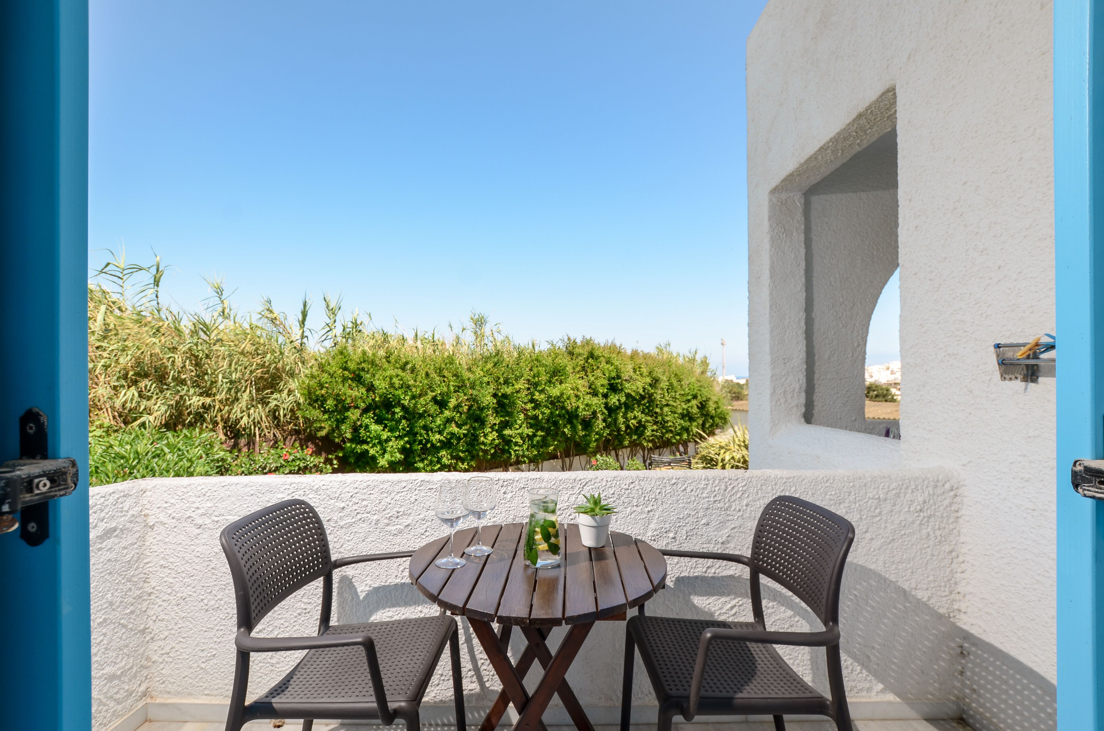 A round wooden table with two chairs on a balcony, a potted plant, and a clear view of a vineyard in the background.