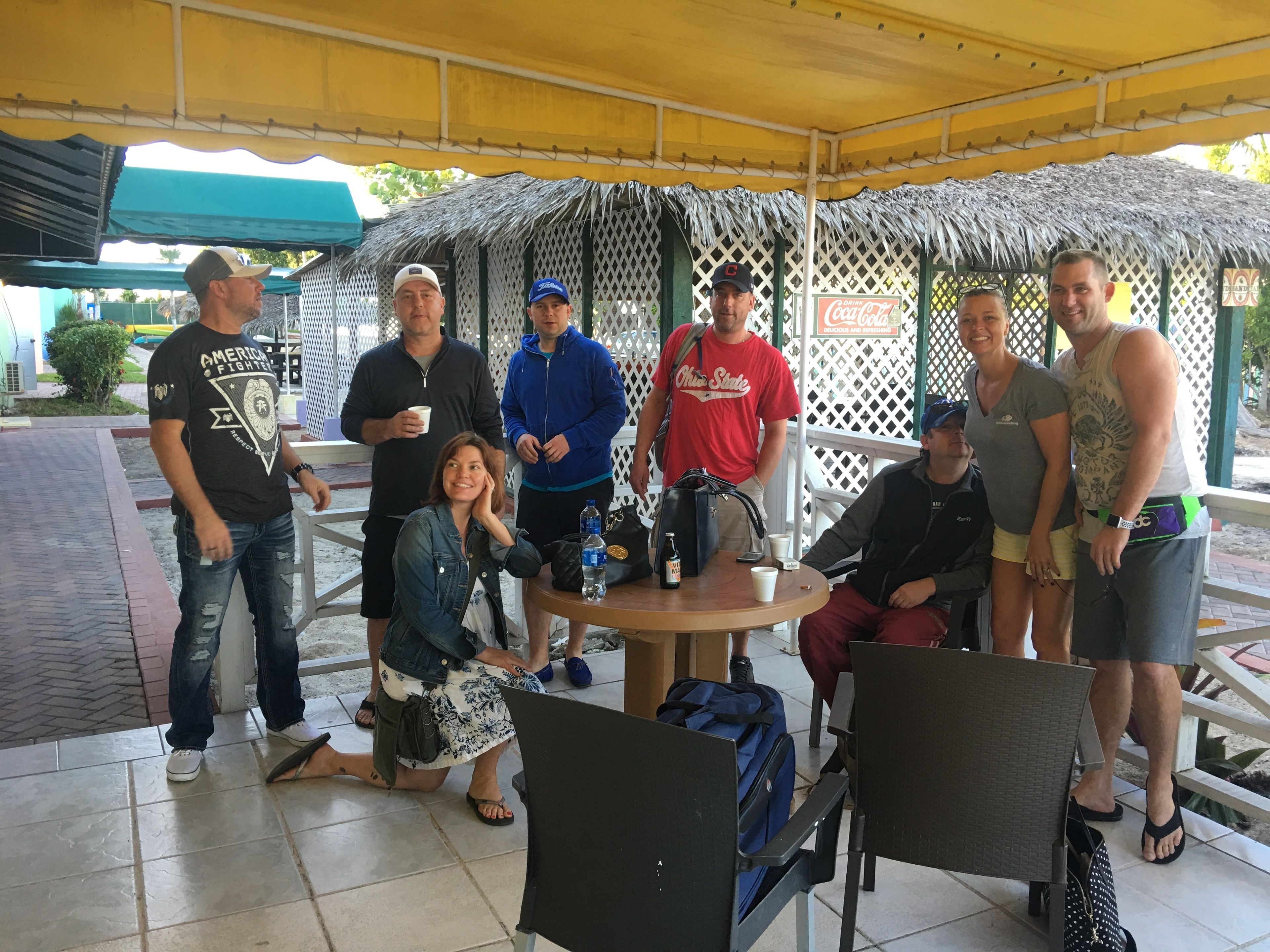 Un grupo de personas reunidas alrededor de una mesa bajo una estructura de techo de paja, con un letrero de Coca-Cola al fondo.